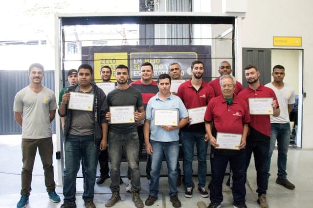 Turma de instaladores credenciados reunida em galpão da Zelo, cada um exibindo o certificado de conclusão, diante do painel da marca e da placa de recepção.